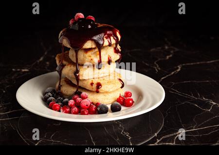 mirtilli, ribes rosso e nero con pancake in una pila, conditi con sciroppo su un piatto bianco su sfondo scuro Foto Stock