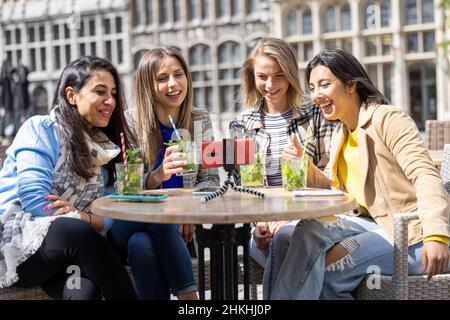 Anversa, Belgio, 21 maggio 2021, quattro turisti o studenti di razza mista seduti fuori nel centro storico della città in una caffetteria terrazza con una videochiamata utilizzando il telefono cellulare. Foto di alta qualità Foto Stock