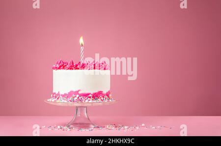 Torta di compleanno con crema di burro rosa, spolverini colorati e candela di compleanno illuminata su sfondo rosa. Foto Stock