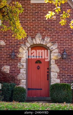 Porta ad arco di legno rosso circondata da una cornice di roccia in una casa di mattoni con castello come ferramenta con foglie caduta appese in giù in un giorno d'autunno Foto Stock