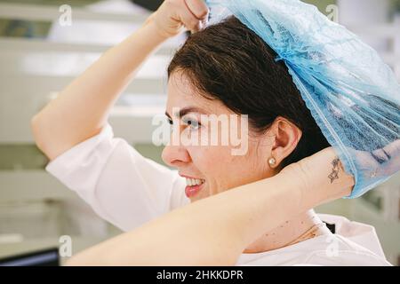 Vista laterale foto di una donna medico sul lavoro mettendo cappuccio medico sulla sua testa con sorriso viso espressione. Foto Stock
