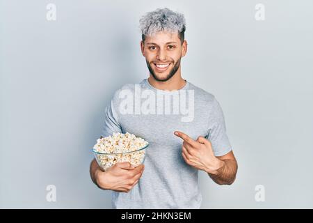 Giovane uomo ispanico con moderni capelli tinti mangiando popcorn sorridendo felice indicando con mano e dito Foto Stock