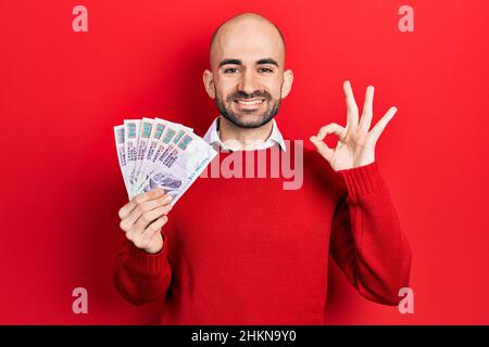 Giovane uomo calvo in possesso di banconote egiziane in sterline facendo ok segno con le dita, sorridente amichevole gesturing eccellente simbolo Foto Stock