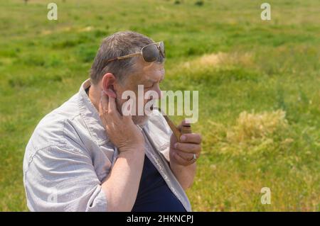 Ritratto con vista laterale del tubo del tabacco caucasico senior fumante mentre si siede all'aperto contro pascolo primaverile Foto Stock