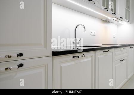 Vista laterale moderna ed elegante cucina con cassetti e armadi bianchi, piano di lavoro in marmo nero e lavello costruito con rubinetto d'argento Foto Stock