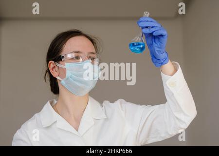 Scienziato donna in guanti blu e occhiali protettivi per analisi liquido blu in fiasca. Foto Stock