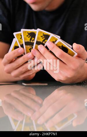 Giovane uomo che gioca una notte Ultimate Werewolf con mano di carte soffiato fuori; famiglia gioco notte a casa; vetro riflettente tavolo da cucina. Foto Stock