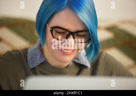 Donna bianca nerdy con capelli blu che lavora su un computer portatile. Felice giovane adulto che fa lavoro freelance sul PC portatile a casa in blocco. Cherfu Foto Stock