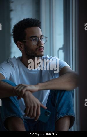 Attraente uomo afroamericano in occhiali e una maglietta bianca si siede su un davanzale. Foto Stock