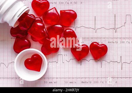 Bottiglia e dispersione di figure sotto forma di cuori rossi sullo sfondo di un cardiogramma (ECG). Il concetto di 'vitamine per un cuore sano' Foto Stock
