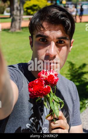 immagine verticale di un giovane latino caucasico ispanico che prende un selfie in un parco pubblico con un fiore in mano Foto Stock