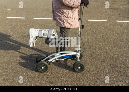 Scooter ginocchia o camminatore che viene utilizzato da donna con piede gamba in stivale chirurgico in giorno d'inverno soleggiato, paesaggio. Foto Stock