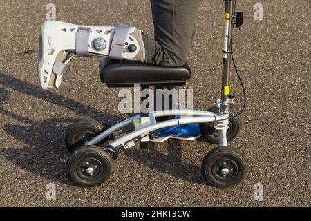 Lo scooter o il camminatore del ginocchio sono usati dalla donna con il piede della gamba in stivale chirurgico nella giornata d'inverno di sole, paesaggio, primo piano. Foto Stock