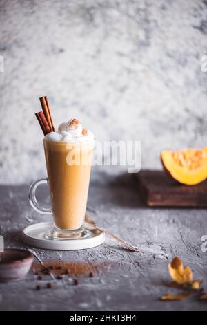 Latte piccante con zucca e panna montata su sfondo grigio. Caffè caldo in tazza di vetro e foglie d'autunno. Ricetta per frullati caldi. Foto Stock