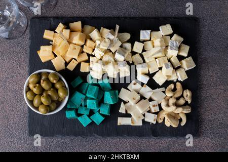 Assortimento di formaggi d'élite a fette con olive e anacardi su piatto d'ardesia su sfondo marrone. Spuntini Wine Party, vista dall'alto Foto Stock