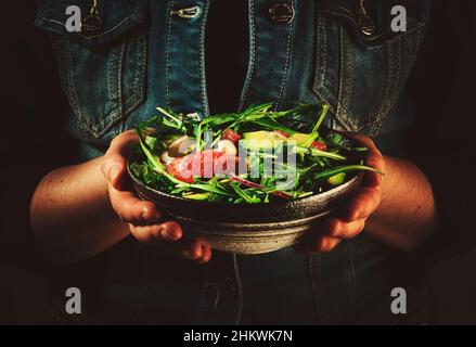 Pranzo vegano sano. Donna in denim blu tiene una ciotola in ceramica con avocado, rucola, pompelmo e insalata di anacardi nelle sue mani. Comfort, pulito, vegetaria Foto Stock