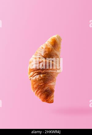 Croissant appena sfornato levitating o volare. Croissant volare in aria isolato su sfondo rosa. Scena di levitazione. Copia spazio per il testo. Cartello mockup. Cibo creativo per la colazione Foto Stock