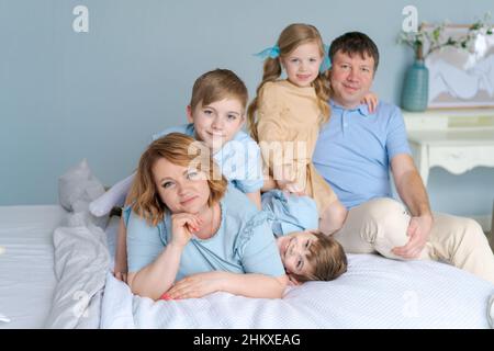 Famiglia allegra che gioca insieme a letto. I genitori trascorrono il loro tempo libero con la loro figlia e due figli. Grande famiglia caucasica che gioca a casa in camera da letto. Mamma e papà stanno ingannando con i bambini Foto Stock