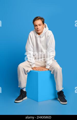 L'uomo allegro in felpa bianca e pantaloni felpa siede su un cubo di legno sullo sfondo blu dello studio Foto Stock