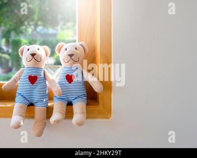 Due allegri orsi bruni sorridenti, bambole in camicie a strisce blu e bianche con cuori rossi, seduti sul bordo di vetro di legno finestra su parete bianca retro gr Foto Stock