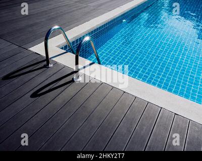 Cornice in pietra sulla piscina quadrata con acqua pulita, nessuno. Piscina a forma quadrata con piastrelle blu con maniglioni scala su legno scuro Foto Stock