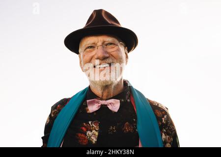 Uomo anziano sorridente mentre si alza all'aperto durante il giorno. Uomo anziano allegro che indossa colorati abiti casual in estate. Spensierato uomo maturo e. Foto Stock