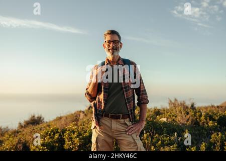 Escursionista maturo che sorride alla macchina fotografica al tramonto. Happy escursionista maturo in piedi sulla cima di una collina mentre si trasporta uno zaino. Avventuroso zaino uomo enjo Foto Stock