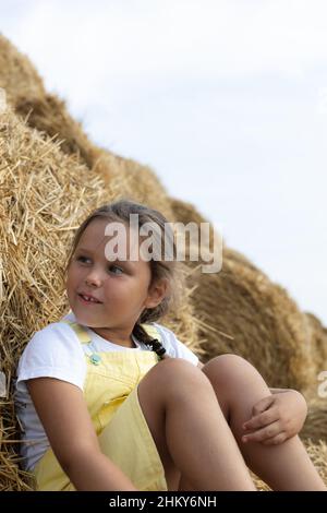 Ritratto di ragazza piccola curiosa seduta su haystack con un altro stack dietro di guardare via con gli occhi interessati con decine di haystack in Foto Stock