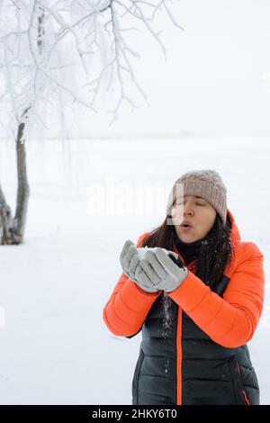 Ritratto di donna splendente di mezza età che soffia la neve dalle mani in guanti con ramoscelli ricoperti di neve sullo sfondo a piedi nella foresta innevata. Magia Foto Stock