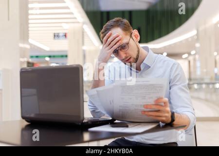 Giovane uomo d'affari perplesso che lavora in un vuoto food Court in centro commerciale con documenti e laptop. Le dichiarazioni fiscali di archiviazione funzionano con i contratti Foto Stock