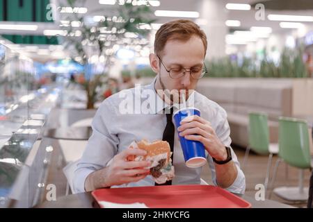 Un impiegato che mangia hamburger fast food e bibita di soda presso un'area ristoro nel centro commerciale a pranzo. Cibo spazzatura al lavoro Foto Stock