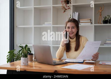 Interessante giovane donna asiatica d'affari che parla sul telefono cellulare in ufficio mentre lavora per il finanziamento con computer portatile e calcolatrice Foto Stock