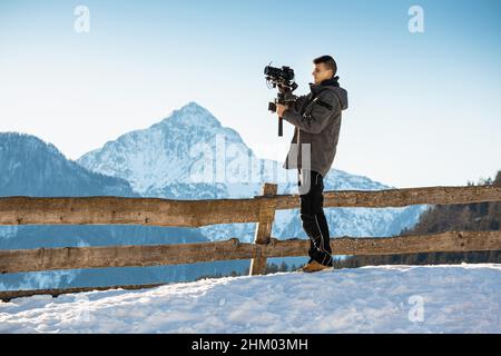 Uomo videografo che realizza riprese utilizzando una fotocamera montata su un dispositivo stabilizzatore gimbal. Neve e montagne sullo sfondo. Foto Stock