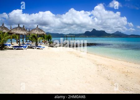 Vista su ombrelloni di paglia e sedie a sdraio su una meravigliosa spiaggia di sabbia bianca e acqua turchese a Mauritius, vulcani che si innalzano sullo sfondo Foto Stock