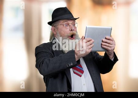 Emotivo scioccato uomo d'affari senior che guarda al tablet pc digitale. Sfocatura dello sfondo interno. Foto Stock