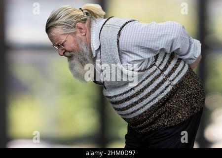 Vista laterale dell'uomo vecchio bearded che soffre dal dolore alla schiena. Sfocatura dello sfondo interno. Anziani e problema di salute concetto. Foto Stock