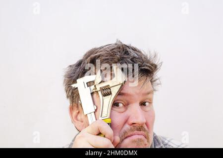 Un maschio adulto stanco mette un attrezzo della mano del metallo alla sua fronte. Contiene una pinza e una chiave. Un uomo con capelli castani aggrovigliati e diseveleati contro un gra chiaro Foto Stock