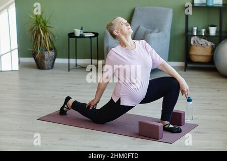 Donna anziana contemporanea in t-shirt e leggins guardando verso l'alto durante la pratica di yoga esercizio sul tappeto in soggiorno Foto Stock