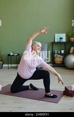 Donna anziana attiva in leggins nero e maglietta beige facendo esercizio fisico su stuoia yoga contro piante domestiche e mobili in soggiorno Foto Stock