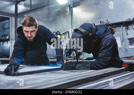 Saldatrice profesionale in uniforme protettiva e maschera di saldatura del telaio in metallo sul tavolo industriale con altri operatori dell'officina industriale. Scatto orizzontale Foto Stock