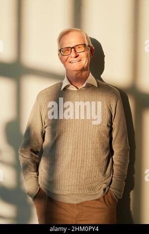 Uomo anziano allegro in pullover beige e pantaloni marroni che si erge soleggiato contro la parete e guarda la macchina fotografica Foto Stock