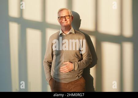 Uomo anziano sorridente in pantaloni marroni e pullover e occhiali in piedi contro la parete illuminata dal sole con ombre e guardando la macchina fotografica Foto Stock