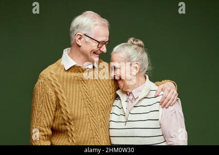 Ridendo coppia senior in abiti casual in piedi su sfondo verde scuro, uomo in pullover abbracciando sua moglie Foto Stock