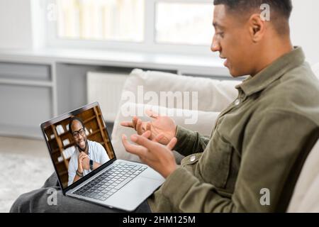 Vista laterale della riunione virtuale dedicata al maschio coinvolto, webinar. Uomo multirazziale che fa videochiamata sul portatile con il suo collega sul divano a casa Foto Stock