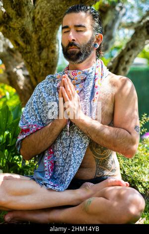 Giovane uomo barbuto svestito praticando yoga (posa di loto) all'esterno in un giardino vicino ad un olivo. Foto Stock