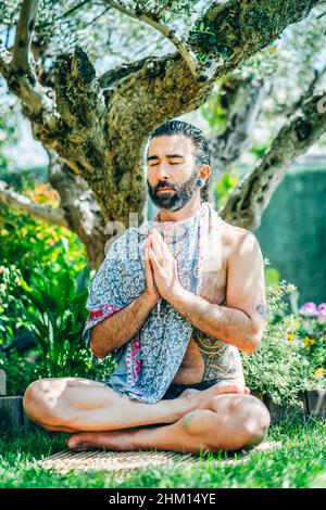 Giovane uomo barbuto svestito praticando yoga (posa di loto) all'esterno in un giardino vicino ad un olivo. Foto Stock