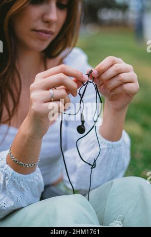 Un teenager striccante filo nero cuffie per ascoltare musica. Lei è seduta sull'erba in un parco mentre cerca di rilassarsi. Foto Stock