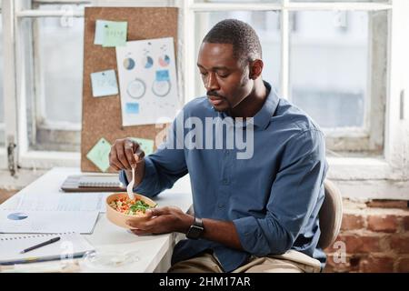 Giovane operaio africano-americano contemporaneo che ha insalata vegetariana fatta in casa per pranzo mentre si siede sul posto di lavoro Foto Stock