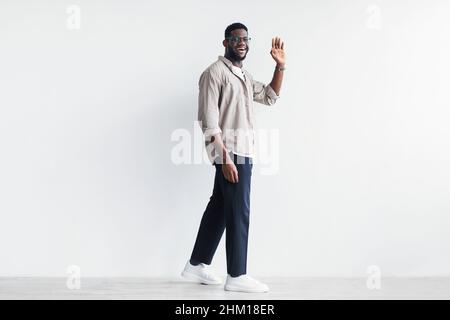 Bel giovane uomo nero che cammina e agitando la mano alla macchina fotografica, salutando qualcuno contro il muro bianco, spazio copia Foto Stock