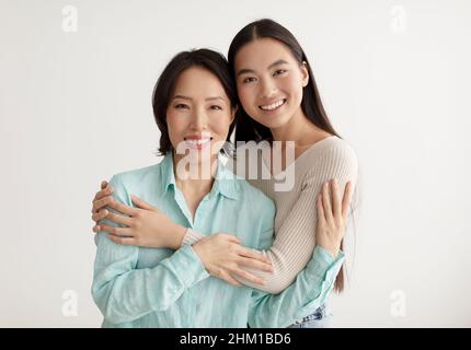 Gioiosa giovane donna asiatica e sua madre anziana che abbraccia e sorride alla macchina fotografica su sfondo bianco studio Foto Stock